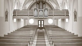 Interior view of a church sanctuary with rows of pews, an organ loft, and arched windows, reflecting the congregation’s fo...