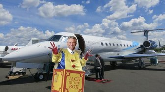 Jack Burrell posing in a cardboard box labeled “Jack in the Box” next to a private jet, referencing controversy within Fai...
