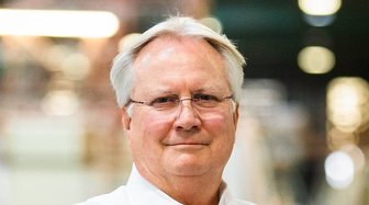 Man with glasses smiles directly at camera; blurred lights in background suggest an office setting